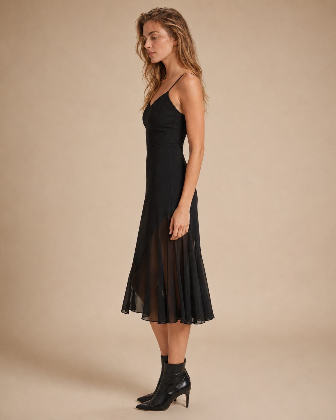 A woman modeling a black spaghetti-strap midi dress with a sheer pleated skirt overlay, paired with black ankle boots, against a solid beige studio background.
