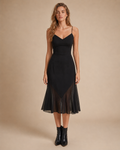 A woman modeling a black midi dress with spaghetti straps, a structured bodice, and a sheer mesh overlay at the skirt, paired with black ankle boots against a warm beige studio background.