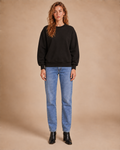 A woman stands against a warm beige studio backdrop wearing a black crewneck sweatshirt, light blue straight-leg jeans, and black ankle boots.