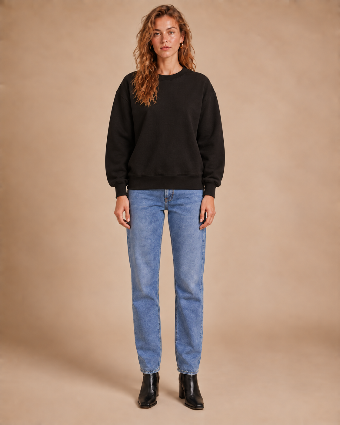 A woman stands against a warm beige studio backdrop wearing a black crewneck sweatshirt, light blue straight-leg jeans, and black ankle boots.