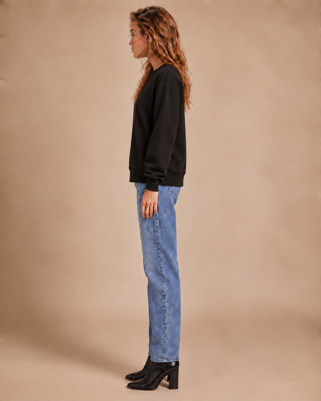 A woman in profile wearing a black crewneck sweatshirt, light-wash blue jeans, and black ankle boots, standing against a solid beige studio backdrop.