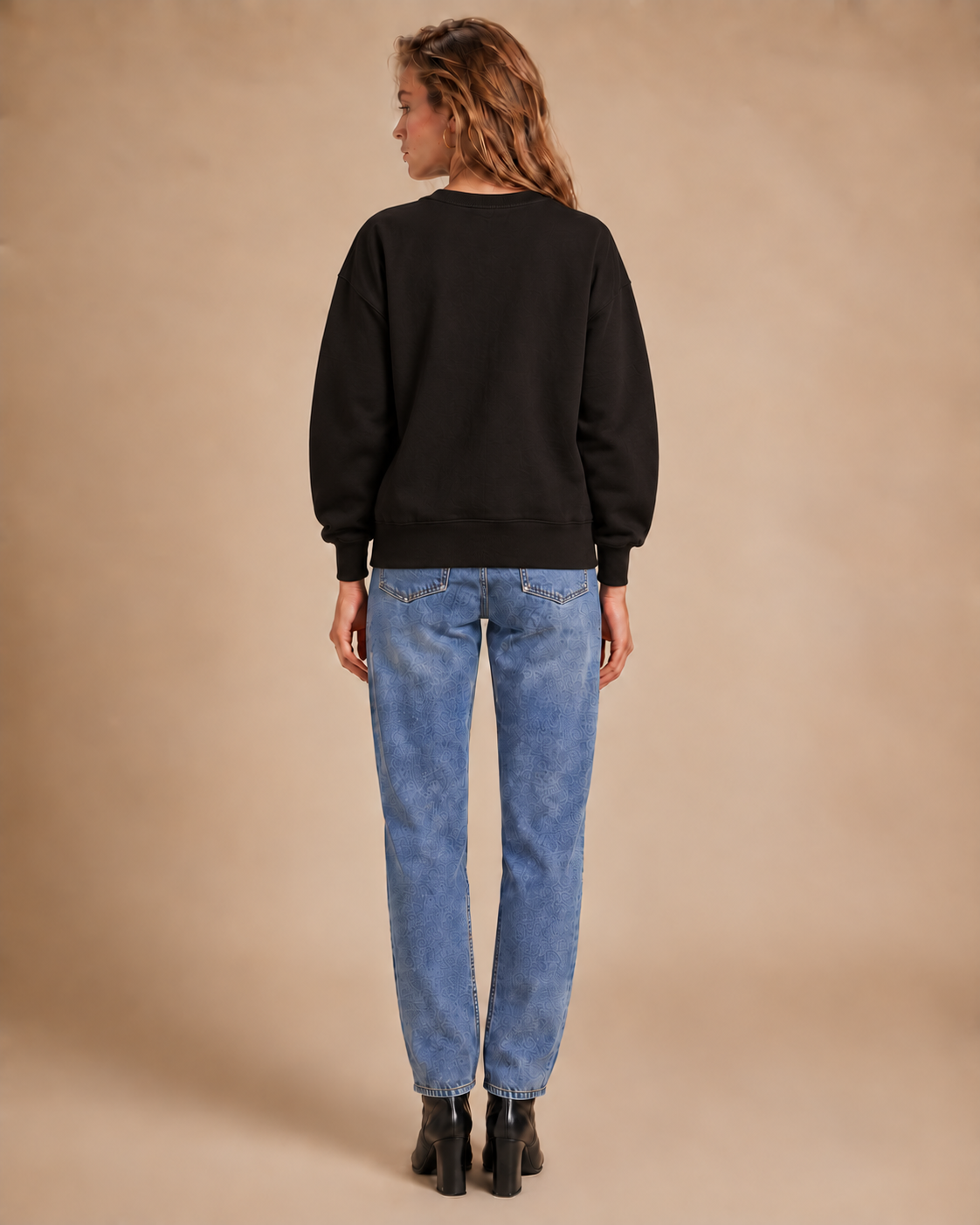 A woman viewed from behind wearing a black crewneck sweatshirt, blue jeans, and black ankle boots, standing against a solid beige studio background.