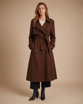 A woman models a long brown trench coat with a tie waist, double-breasted front, and large front pockets, paired with black cropped pants and black ankle boots, against a solid beige studio background.