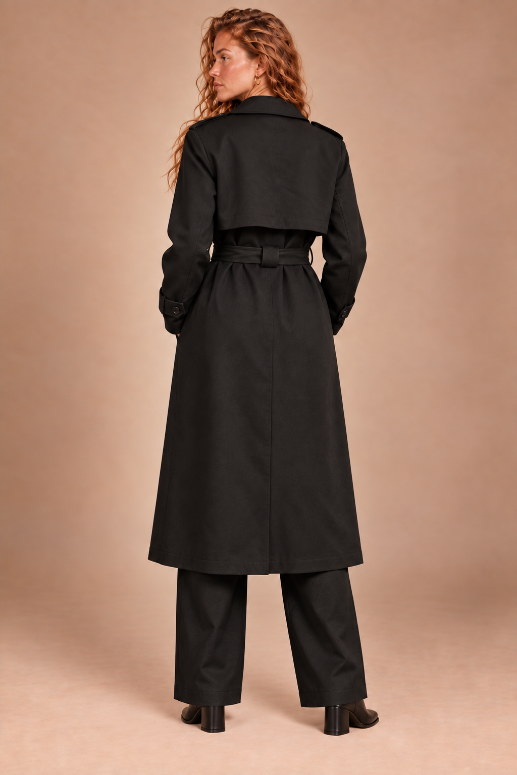 A woman shown from the back wearing a long black trench coat with a matching belt, black wide-leg trousers, and black heels, set against a solid warm beige studio background.