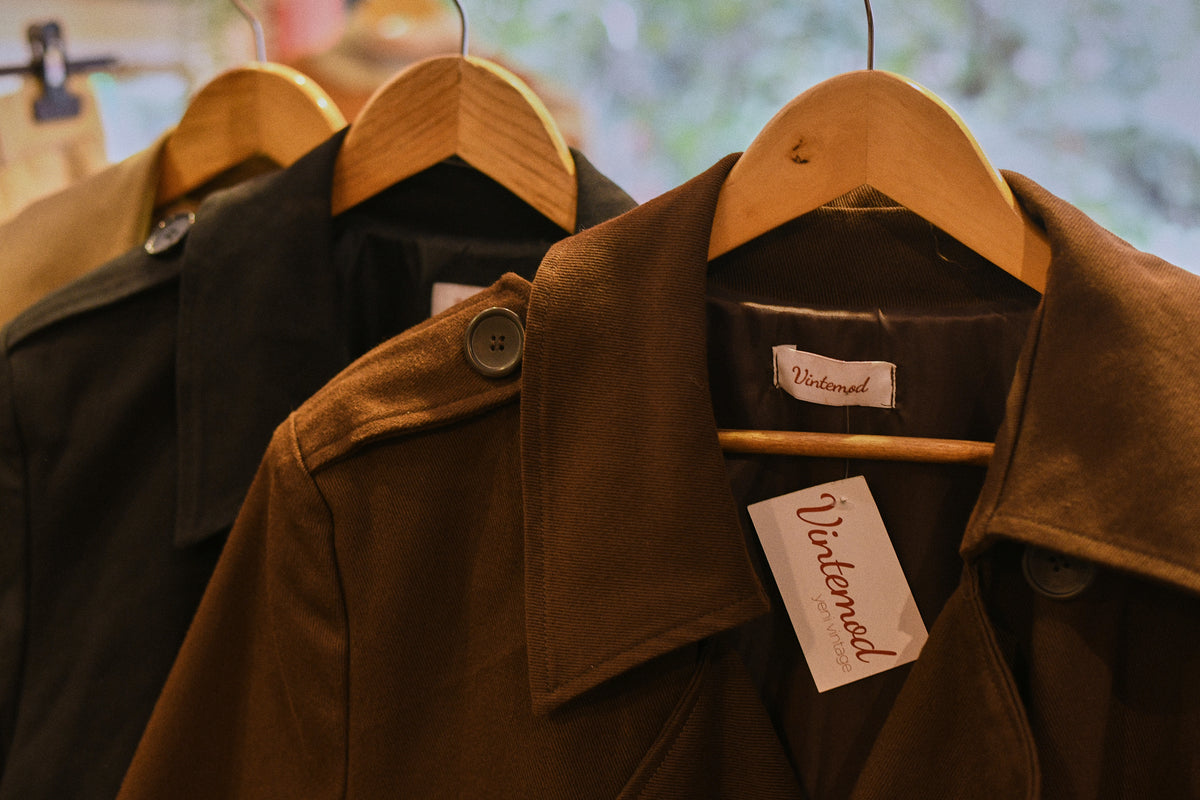 A brown wool-blend trench coat hanging on a wooden hanger, with a white 'Waterfowl' brand tag visible. Another dark coat is partially visible behind it on a wooden hanger.